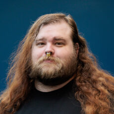 A headshot of a person with curly brown hair, a beard, and mustache.