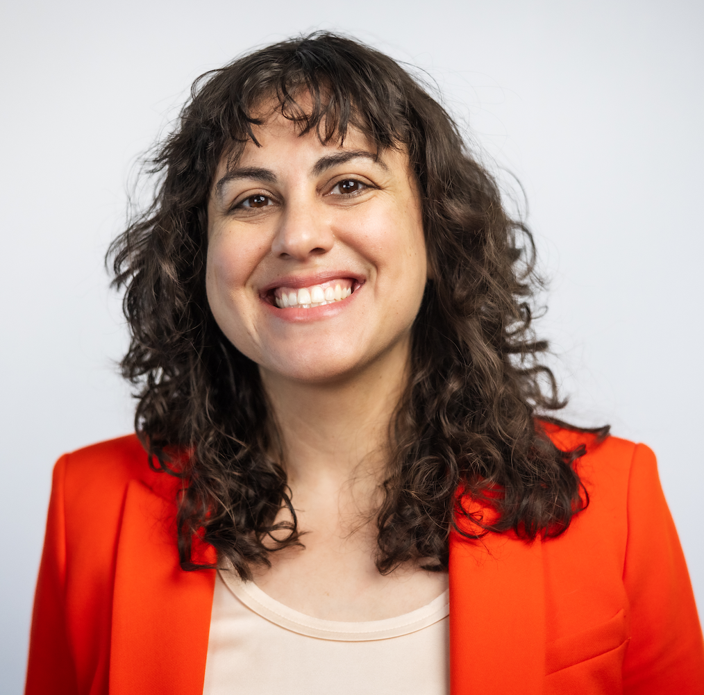 A headshot of a woman in an red orange blazer and cream shirt. 