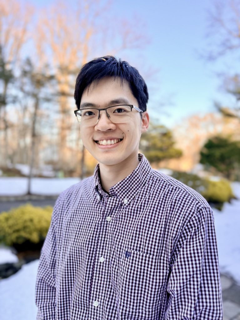 An Asian man with dark hair and glasses in a plaid button down shirt. He's standing on a walkway lined with trees and bushes.