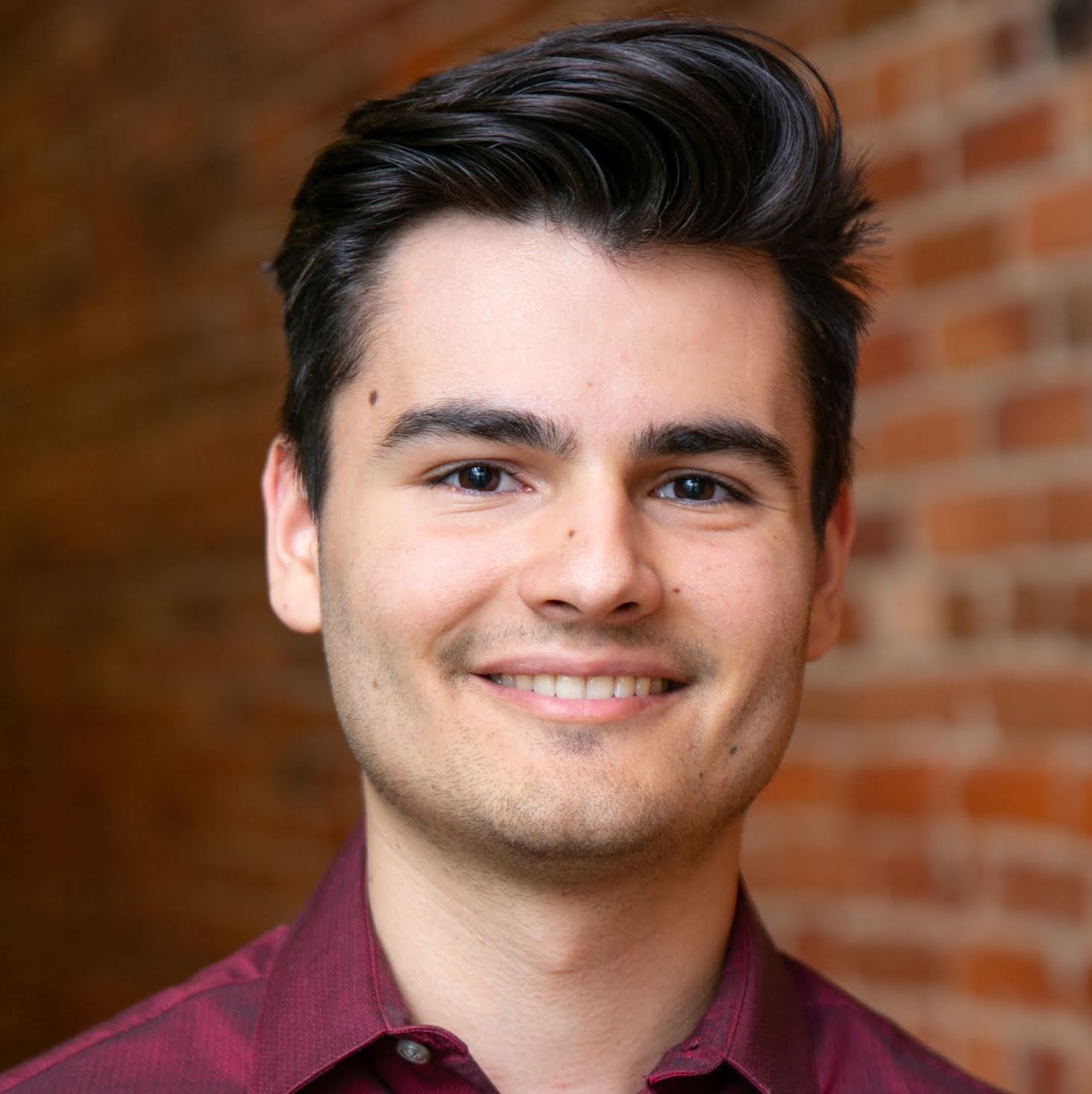 A smiling man with short dark hair in front of a brick wall. He is wearing a burgundy button down shirt.