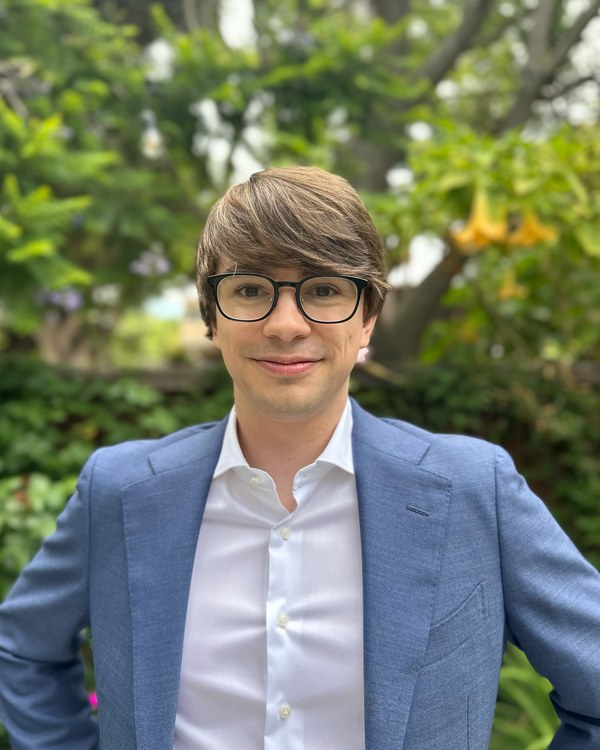 A man with brown hair and glasses standing in front of some foliage. They are wearing a blue blazer and white button down.